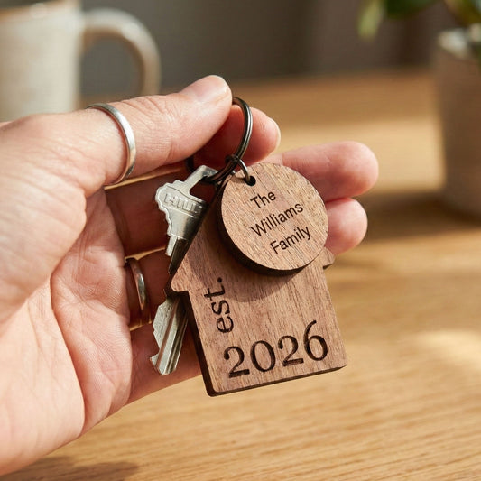 A hand holding a house-shaped wooden keychain engraved with a family name and a year, attached to a metal keyring