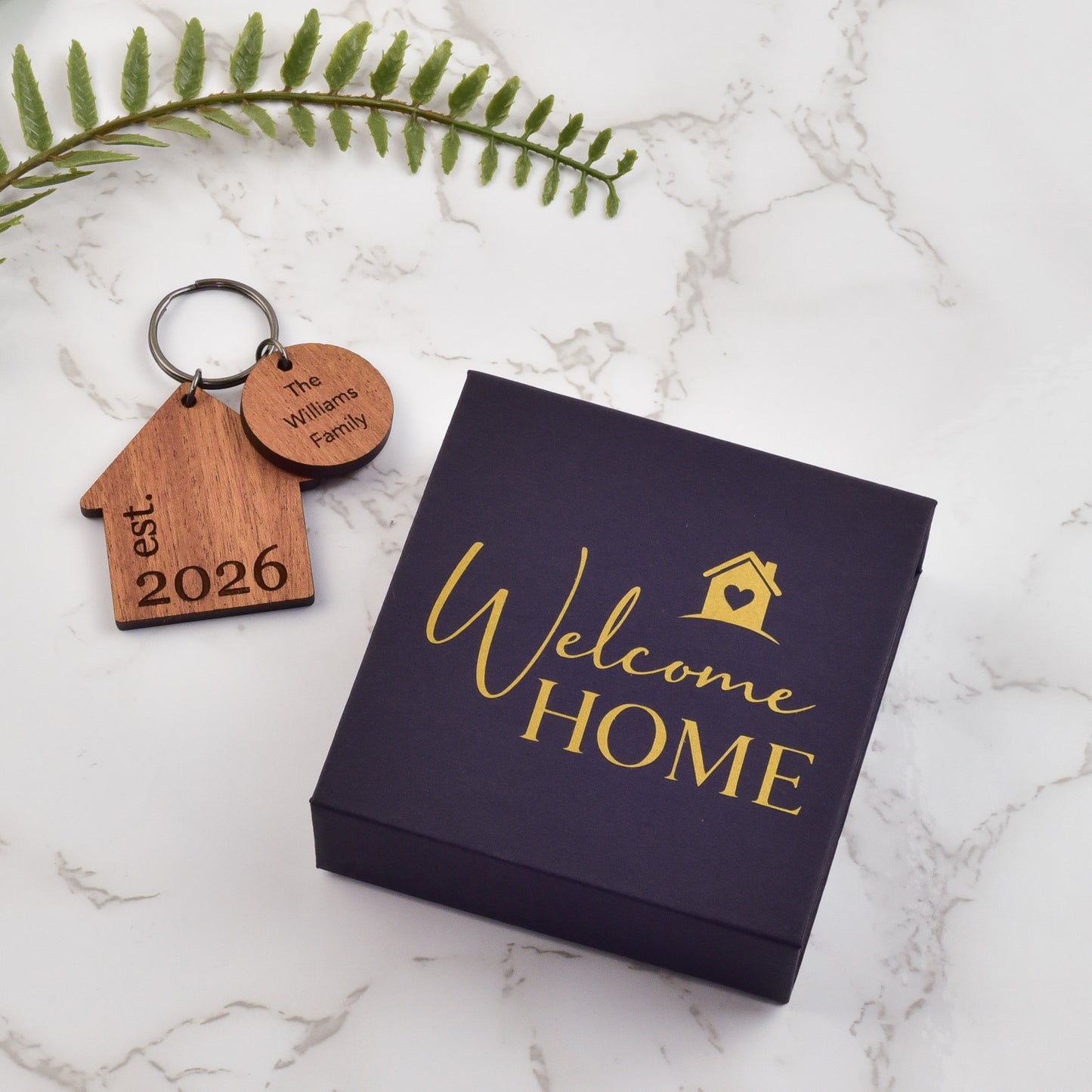 Wooden keychain with engraved text on a 'Welcome Home' card with gold text, placed on a marble surface. 